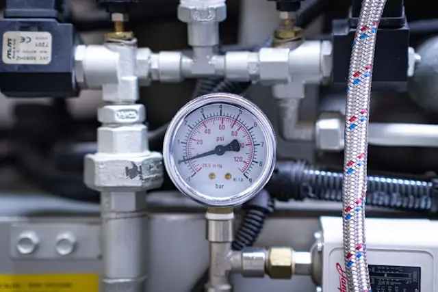 Analoges Manometer mit Bar- und Psi-Skala zwischen Rohrleitungen, Ventilen und einem Druckschlauch an einer Industrieanlage.