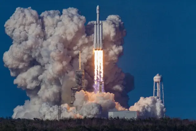 Vom Startkomplex abhebende Rakete mit hellem Triebwerksfeuer und mächtigen Rauchwolken vor blauem Himmel, im Bezug zum SpaceX-Börsengan