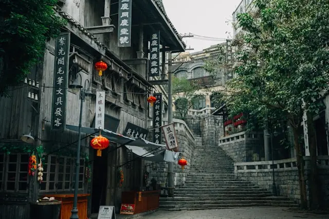 Gasse einer Altstadt mit roten Laternen und chinesischen Schriftzeichen auf Holzfassaden vor einer Steintreppe, ohne erkennbaren Bezug zur Maase-Aktie
