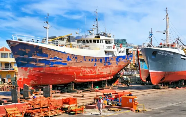 Zwei Fischtrawler mit verwittertem Rumpf in Blau, Rot und Weiß stehen in einem Trockendock einer Werft, umgeben von Werftgebäuden und Arbeitern.