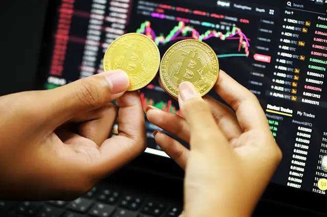  Zwei goldene Bitcoin-Münzen werden vor einem Laptop mit Kryptowährungs-Handelschart gehalten, im Kontext des Börsengangs von Gemini Space Station.