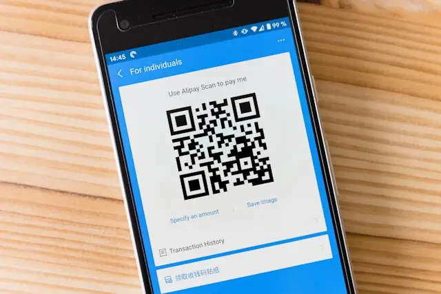 Smartphone zeigt die Bezahl-App Alipay mit QR-Code zum Scannen, aufliegend auf einer Holzoberfläche im Kontext mobiler Zahlungsdienste.