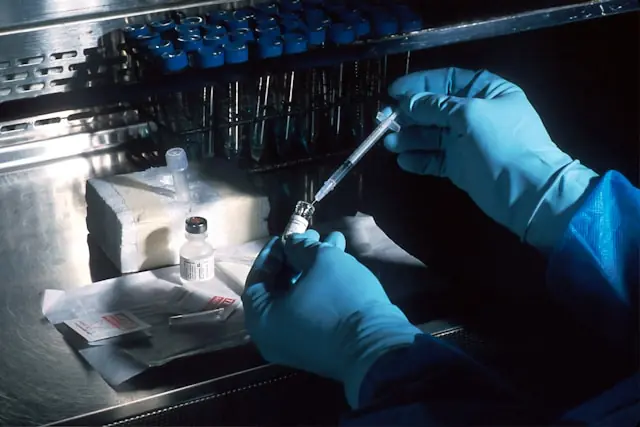 Behandschuhte Hände ziehen mit einer Spritze Flüssigkeit aus einem Fläschchen, im Hintergrund Probenständer, als Bezug zum Biotech-Wert SL Science
