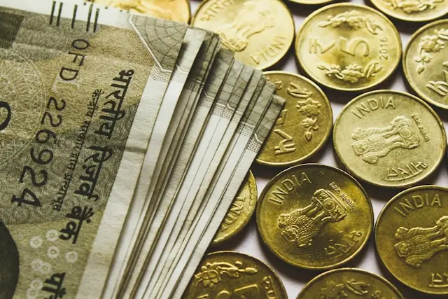 Gestapelte indische Rupien-Banknoten liegen neben goldfarbenen Münzen mit Ashoka-Symbol auf heller Fläche