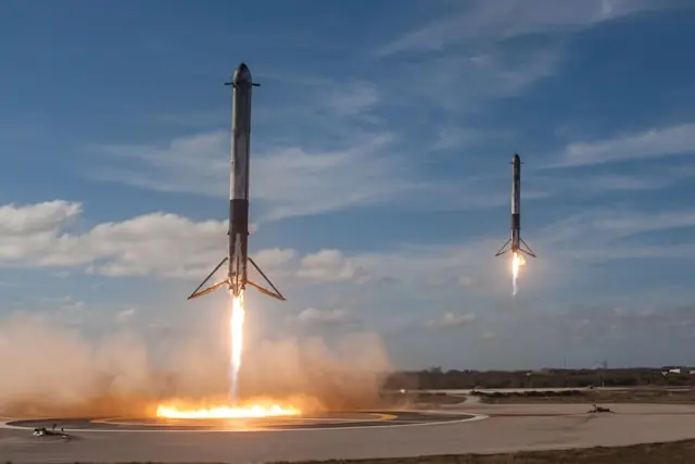 Zwei Raketenstufen mit ausgefahrenen Landebeinen und brennenden Triebwerken bei der Landung auf einer Landefläche unter blauem Himmel, im Bezug zum SpaceX-Börsengang