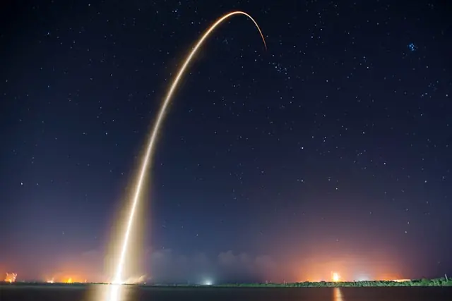 Leuchtende Flugbahn einer startenden Rakete als langer Lichtbogen am Sternenhimmel über der Küste, im Bezug zum überzeichneten SpaceX-Börsengang