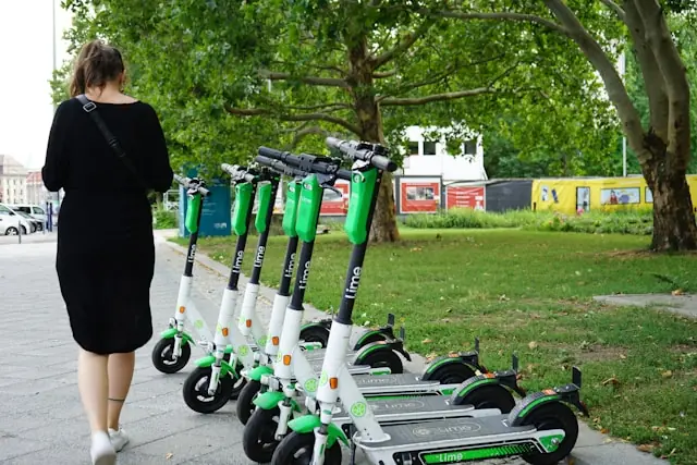 Mehrere grün-weiße Lime-E-Scooter stehen aufgereiht auf einem Gehweg, während eine Frau im schwarzen Kleid vorbeigeht.